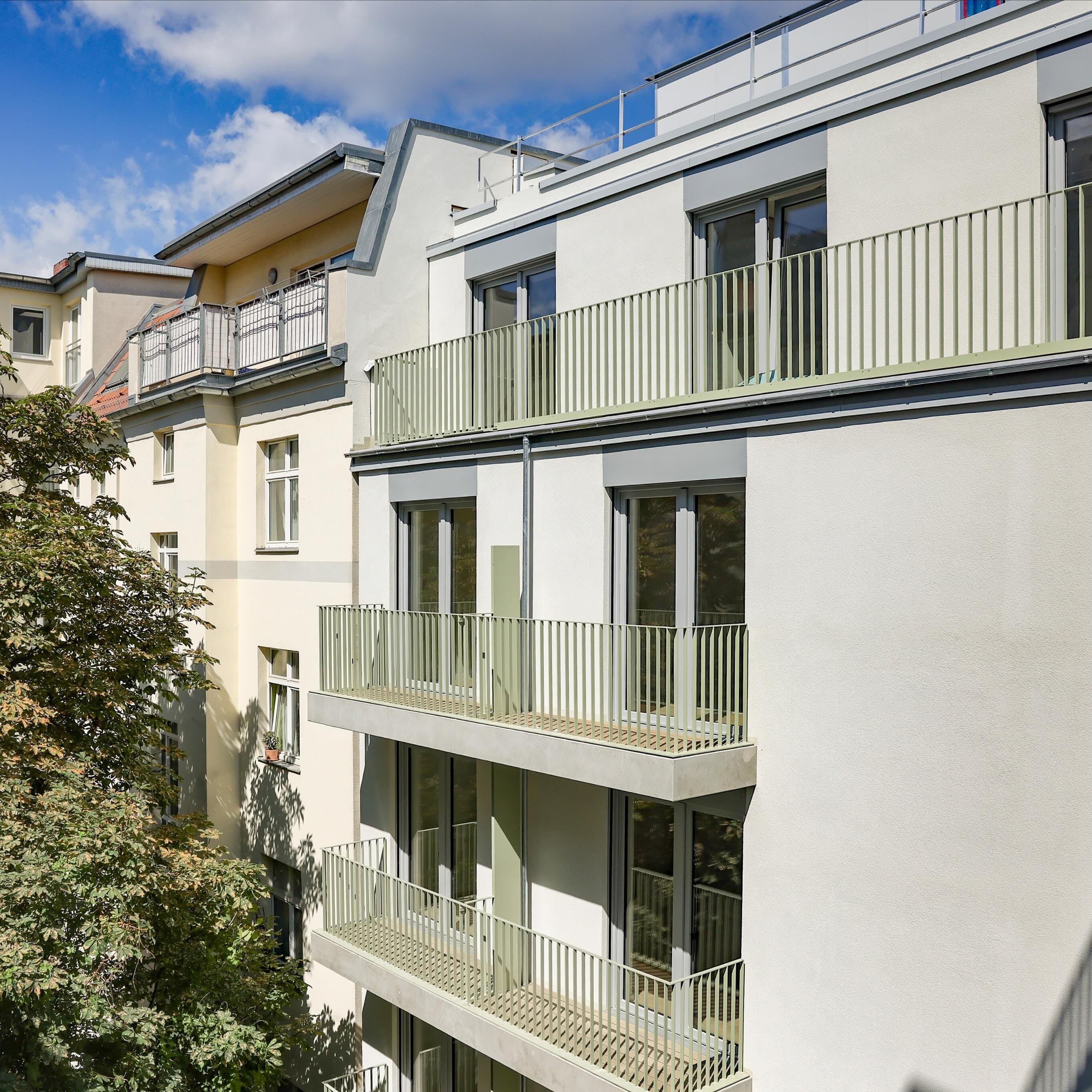 Hofansicht des Apartmenthauses – moderne helle Fassade mit lindgrünen Balkongeländern über mehrere Etagen, daneben Altbauten und ein großer Baum unter blauem Himmel
