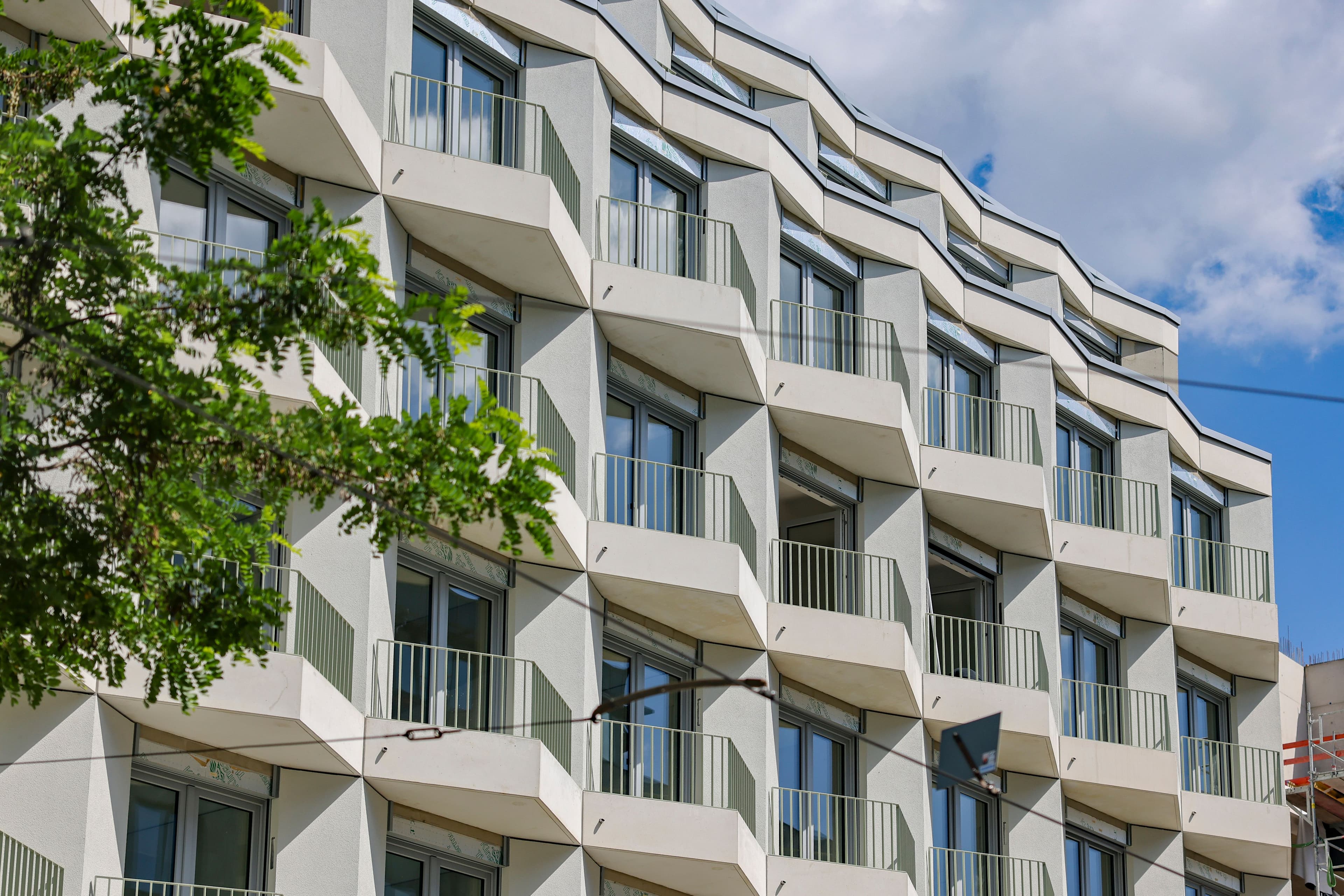 Straßenfassade von FRIEDRICH in Berlin-Friedrichshain: helle, gefaltete Neubaufassade mit versetzten Balkonen und grünen Geländern, davor Baumlaub und der Fahrdraht der Tram.