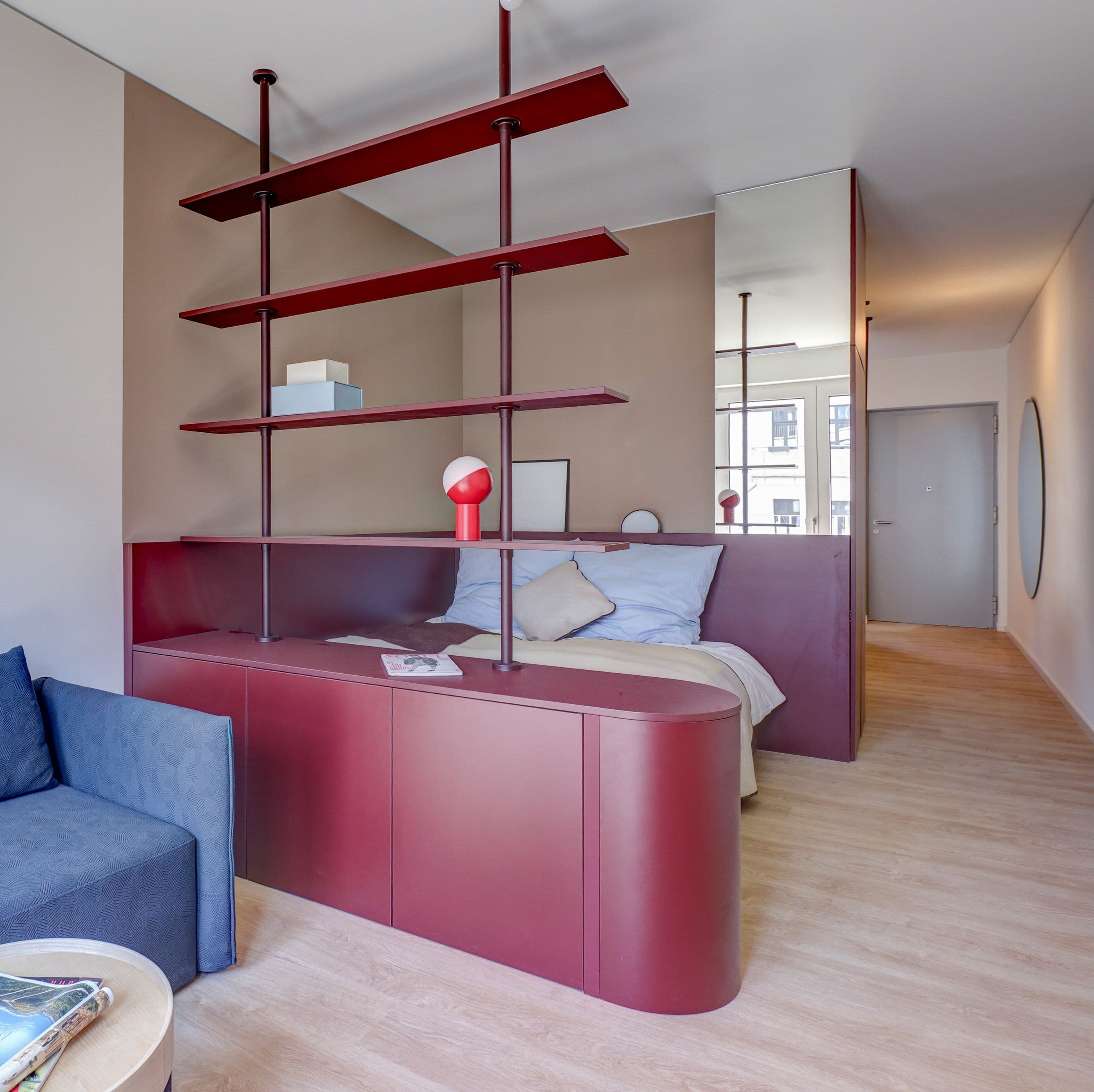 Studio Plus – weinrotes Raumteiler-Regal mit geschwungenem Sideboard trennt das Bett mit hellblauer Bettwäsche vom Wohnbereich mit blauem Sofa