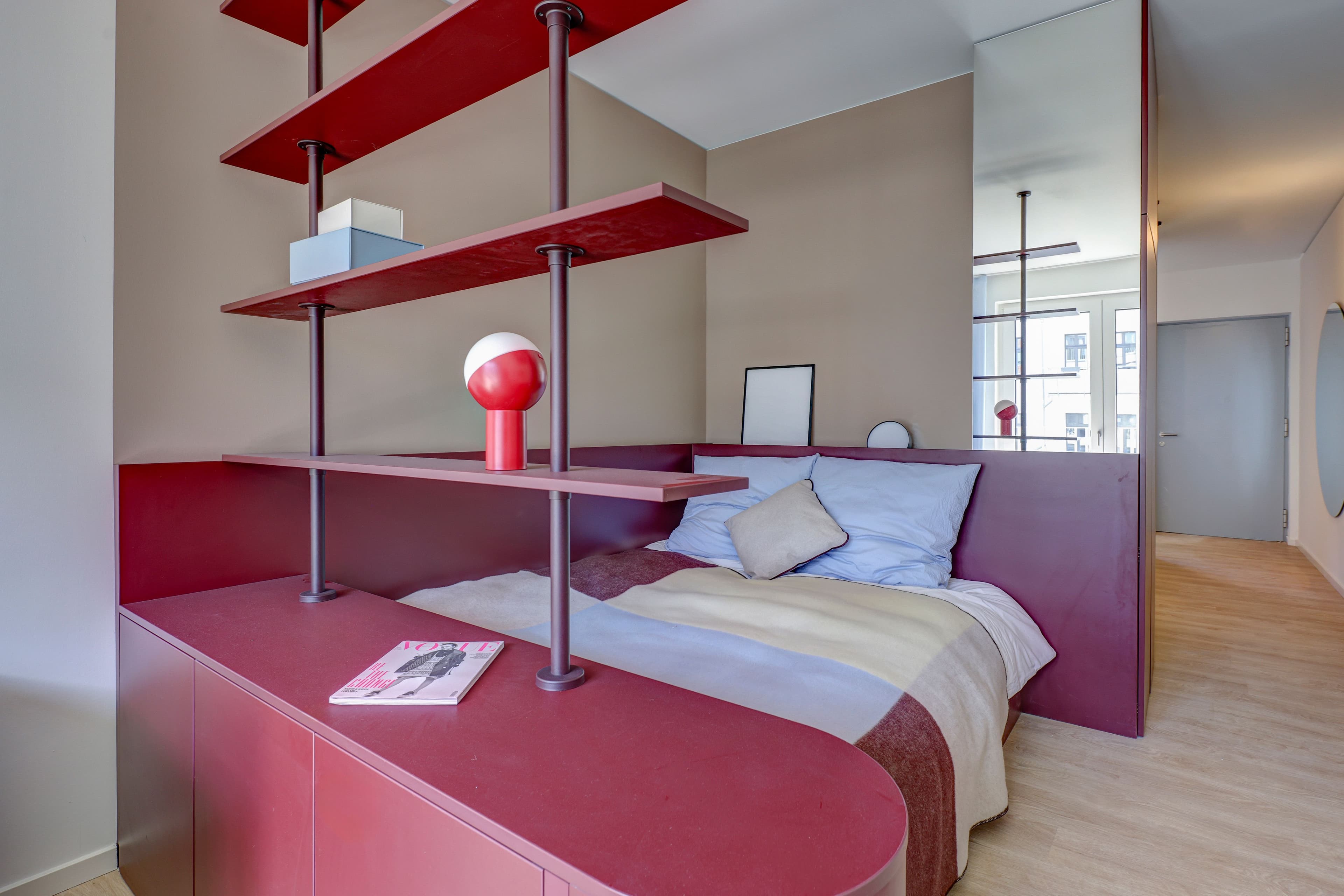 Schlafbereich eines FRIEDRICH-Apartments: Bett hinter einem beerenroten Raumtrennregal mit Sideboard und Tischleuchte, dahinter der Blick in den angrenzenden Flur.